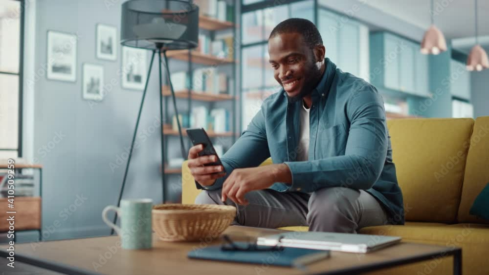 Poster handsome black african american man using smartphone while sitting on a sofa in cozy living room. fr