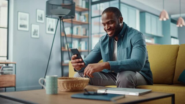Handsome Black African American Man Using Smartphone while Sitting on a Sofa in Cozy Living Room. Freelancer Working From Home. Browsing Internet, Using Social Networks, Having Fun in Flat.