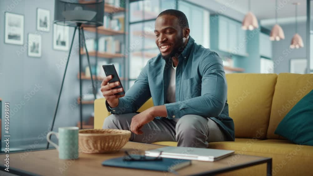 Poster excited black african american man having a video call on smartphone while sitting on a sofa in livi