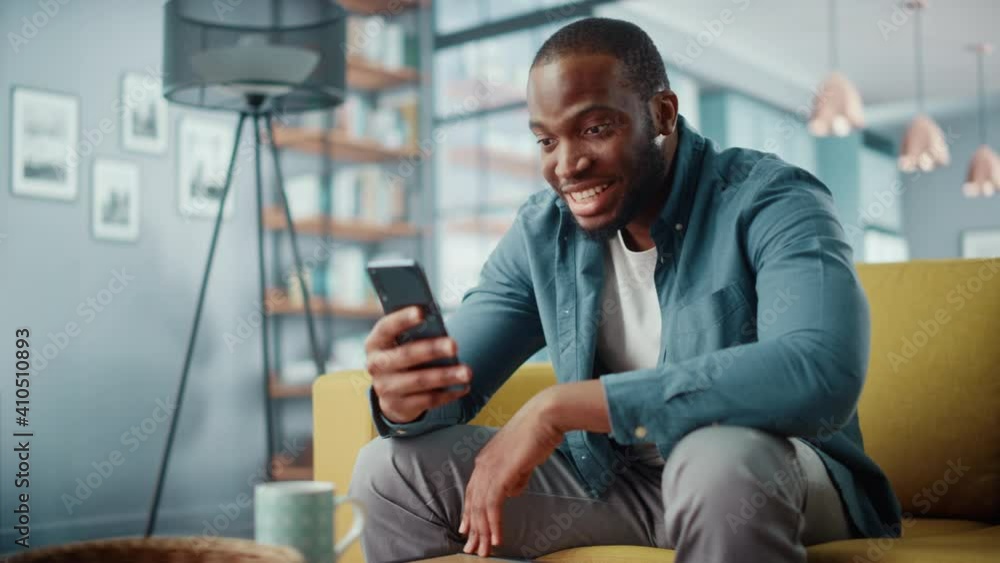 Poster excited black african american man using smartphone while sitting on a sofa in living room. happy ma