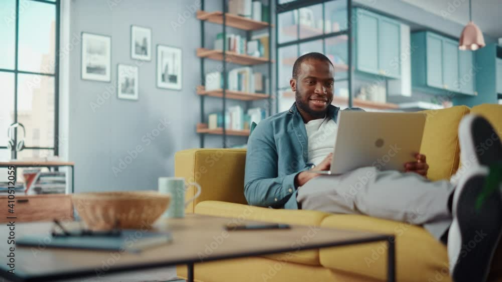 Poster Handsome Black African American Man Working on Laptop Computer while Lying on a Sofa in Cozy Living Room. Freelancer Working From Home. Browsing Internet, Using Social Networks, Having Fun in Flat.