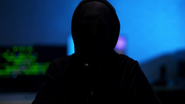Hacker taking off mask in blue room indoors. Unrecognizable man in black hoodie posing. Hacking and coding concept.