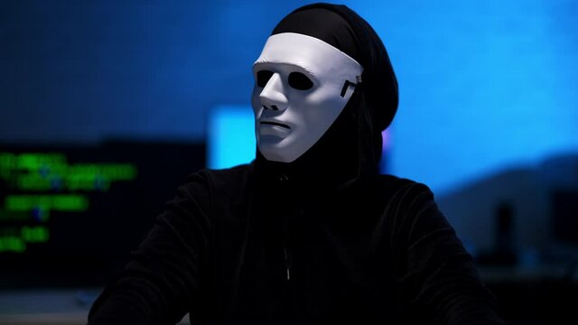 Middle shot of man in black hoodie and anonymous white mask looking around in blue room and typing on keyboard. Unrecognizable hacker in darkness indoors. Hacking and coding.