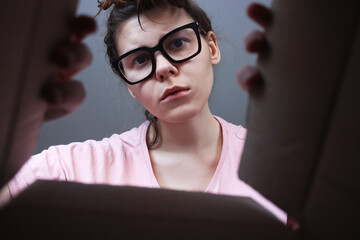 A young woman wearing large medical glasses looks into a box with a bewildered face. Unexpected contents of the package. Unscheduled postal parcel.  Home delivery, falls short of expectations