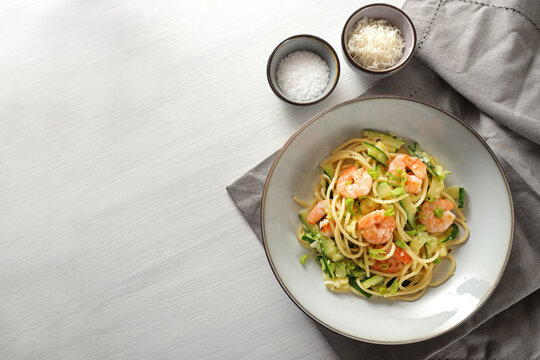 Spaghetti with zucchini courgettes, spring onions and prawns in a grey plate on a white painted table, napkin and small salt and parmesan bowls, copy space, high angle view from above