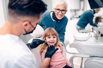 Obraz premium Cute little girl sitting on dental chair and having dental treatment. Her grandmother is with her. Dentist is wearing protective face mask due to coronavirus pandemic.