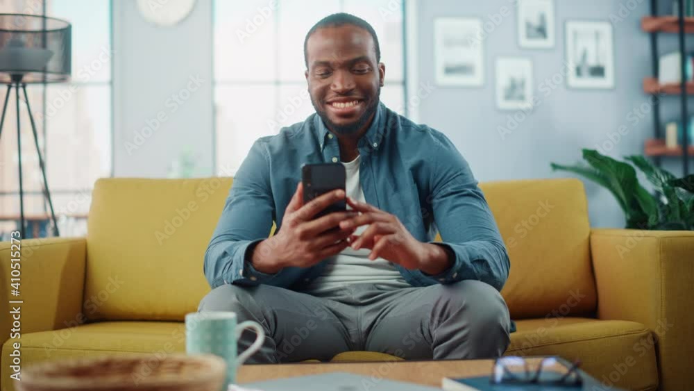Poster excited black african american man using smartphone while resting on a sofa in living room. happy ma