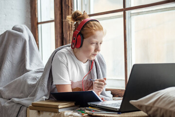 A teenage girl with red curly hair enjoys listening to music sitting on the windowsill. Online training, listens to lectures