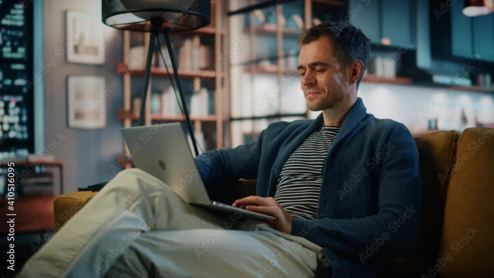Poster Handsome Caucasian Man Working on Laptop Computer while Sitting on a Sofa in Stylish Cozy Living Room. Freelancer Working From Home. Browsing Internet, Using Social Networks, Having Fun in Flat.