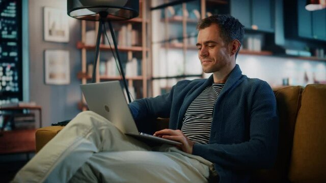 Handsome Caucasian Man Working on Laptop Computer while Sitting on a Sofa in Stylish Cozy Living Room. Freelancer Working From Home. Browsing Internet, Using Social Networks, Having Fun in Flat.
