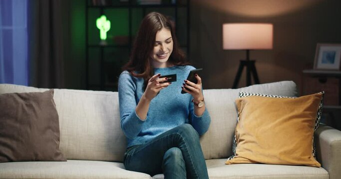 Young happy beautiful woman sitting in evening at home on couch and holding smartphone and bank card rejoices in successful purchase.