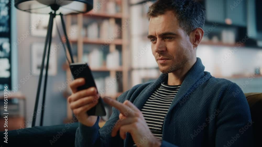 Aufkleber Excited Caucasian Man Using Smartphone while Resting on a Sofa in Stylish Living Room. Happy Man Smiling at Home and Chatting to Colleagues and Clients Over the Internet. Using Social Networks.
