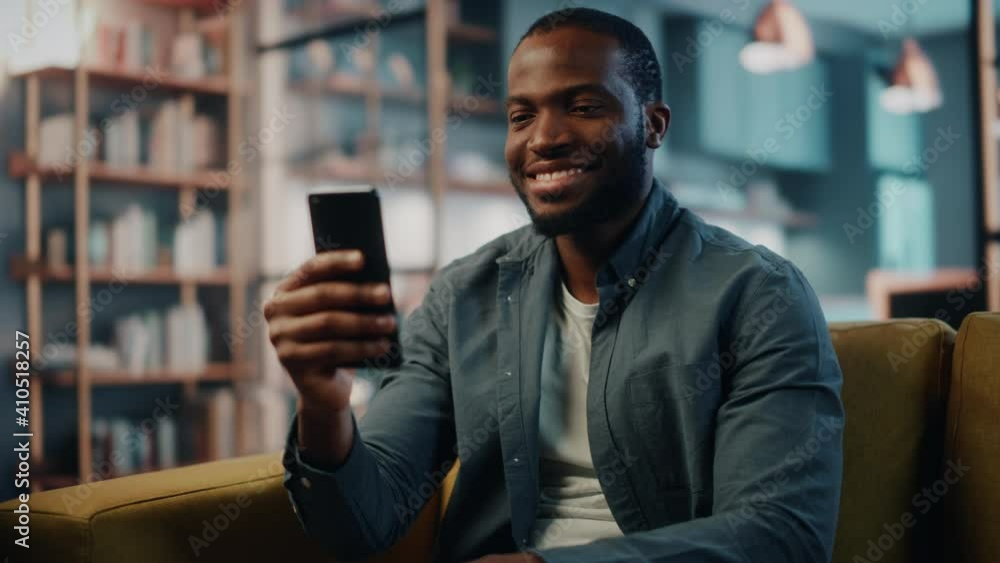 Poster excited black african american man using smartphone while resting on a sofa in living room. happy ma
