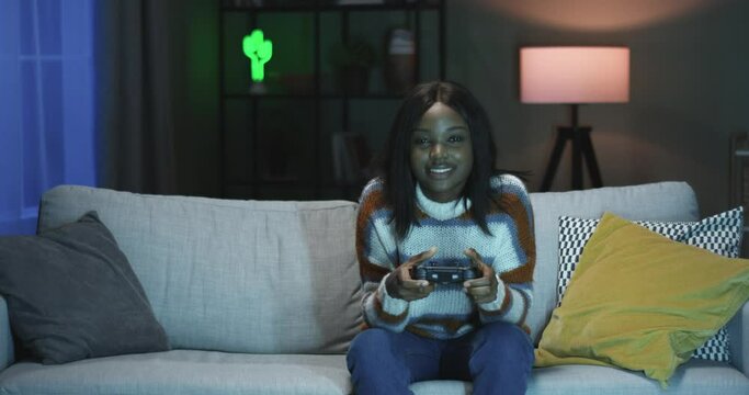 Young happy beautiful African American woman has fun in evening at home playing video games on console.