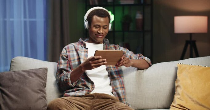 Young handsome african american guy sitting at home on couch at night and watching videos on tablet with headphones.