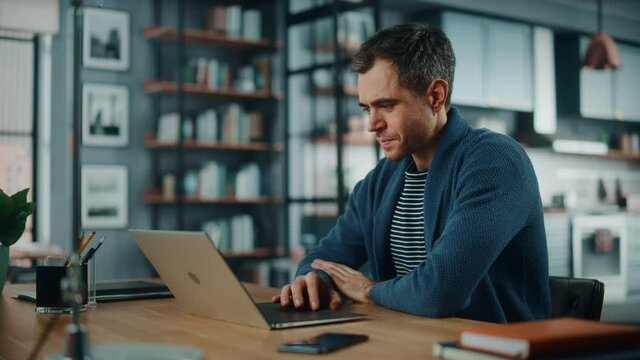 Handsome Caucasian Man Working on Laptop Computer while Sitting Behind Desk in Cozy Living Room. Freelancer Working From Home. Browsing Internet, Using Social Networks, Having Fun in Flat.