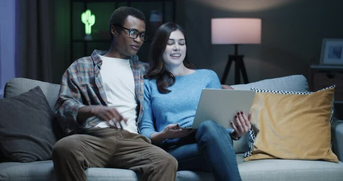 Young happy multinational couple sitting at night on couch at home and talking to friends on video call using laptop and waving hands.