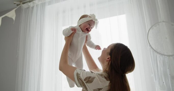 Mother and child have fun at home together. Young beautiful mom throws her adorable newborn baby up and catches. Concept of childhood, new life, parenthood.