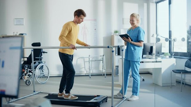 Hospital Physical Therapy Room: Patient with Injury Walking on Treadmill Holding for Parallel Bars, Professional Physiotherapist Assists, Helps, Trains Disabled Person Do Rehabilitative Physiotherapy 