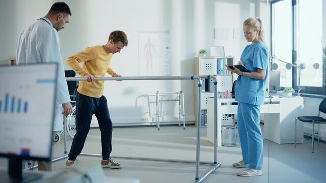 Hospital Physical Therapy Room: Patient with Injury Walking Holding for Parallel Bars, Professional Physiotherapist and Doctor Assist, Help, Train Disabled Person Do Rehabilitative Physiotherapy 