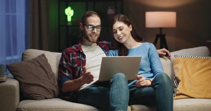 Young happy caucasian couple sitting in evening on couch and talking with family online using web camera on laptop.