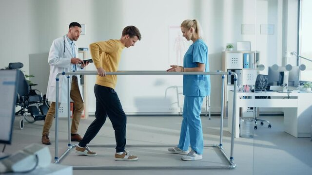 Hospital Physical Therapy: Strong Patient with Injury Making First Steps, Walks Holding for Parallel Bars. Physiotherapist, Rehabilitation Doctor Assist, Help Disabled Person. Side View Slow Motion