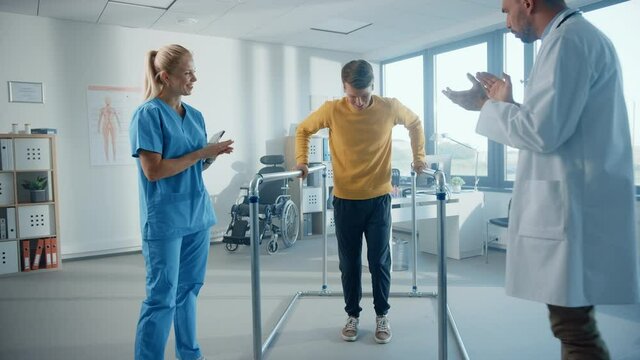 Hospital Physical Therapy: Determined Male Patient with Injury Successfully Walks His First Steps Holding Parallel Bars. Physiotherapist, Rehabilitation Doctor Encourage, Applaud Disabled Person