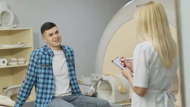 Woman doctor radiologist has a dialogue with young male patient, asks and notes the patient's medical history, complaints in modern clinic beside modern closed-type MRI