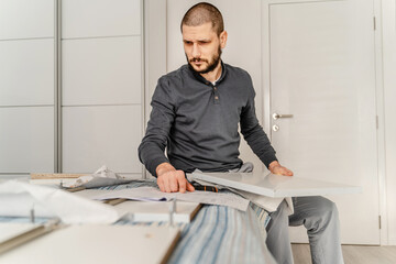 One Man with beard alone sitting while Putting Together Self Assembly Furniture at Home holding...