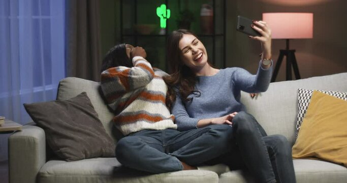 Two best female friends sit on couch in evening and have fun recording joint video for social networks on smartphone.