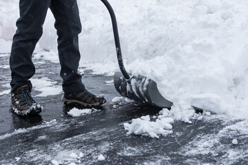 Snow being shoveled off a black asfalt driveway with shovel; person wearing gray pants and boots; big pile of snow