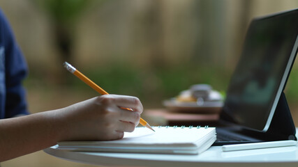 Hand holding pencil to write on blank notebook while online studying in garden at home