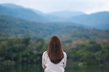 Rear view image of a woman sitting alone by the lake looking at the mountains with green nature background