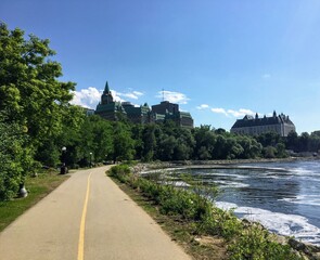 A beautiful bike and walking path along the ottawa river on a sunny summer day with blue sky.  The Supreme Court of Canada is in the distance, in Ottawa, Ontario, Canada. - obrazy, fototapety, plakaty
