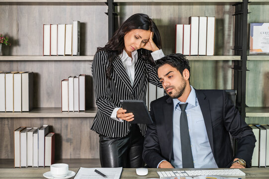 A young businesswoman in a suit showing a tablet screen to a businessman sitting on his desk in an office looking disappointed, unhappy, and worry.
