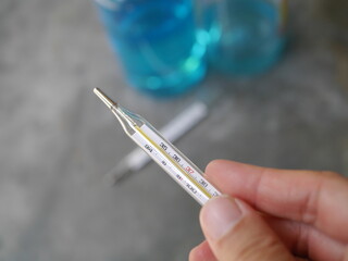 Hand held a close-up of a glass thermometer. Behind there is a picture of hand washing alcohol...