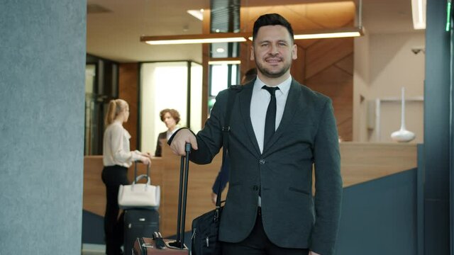 Slow motion portrait of handsome middle-aged businessman in suit standing in hotel lobby smiling looking at camera holding luggage. Reception desk is in background.