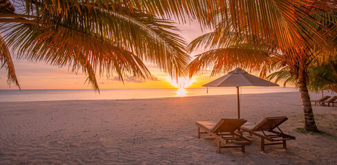 Beautiful tropical sunset scenery, two sun beds, loungers, umbrella under palm tree. White sand, sea view with horizon, colorful twilight sky, calmness and relaxation. Inspirational beach resort hotel