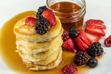 Tasty, yummy breakfast of pancakes stacked with  strawberries, raspberries and black berries placed on a white plate surrounded by maple syrup, honey. Taken in professional studio setting.  