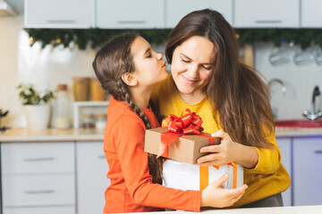 Happy family: daughter kisses her mother a gives gift and flowers for the holiday. A cute girl in the kitchen gives her mom a box and hugs for Mothers day
