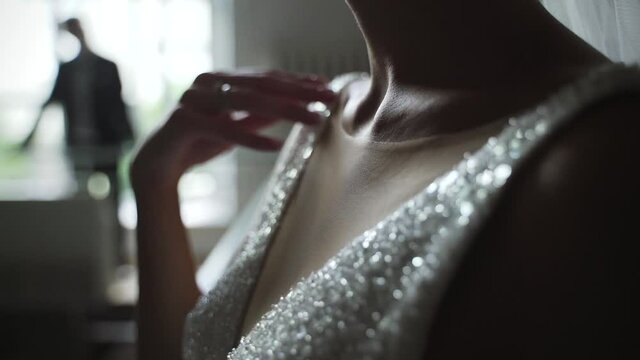Bride in sparkly wedding dress touches her collarbone with her hand. Blurred silhouette of groom in the background 