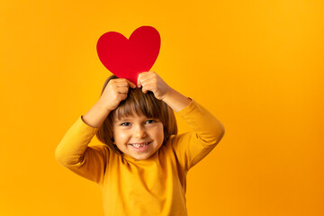 A kid holds a red heart on a yellow background. Little cute girl holds a greeting card in his hand