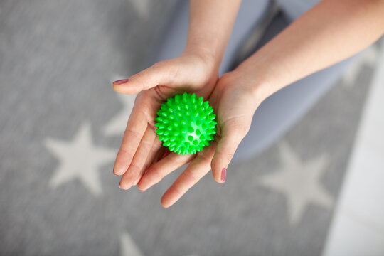 close-up two women's hands hold a green spiked massage ball against a gray carpet with white stars and knees in gray leggings.