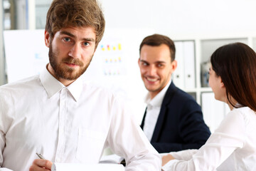 Fototapeta premium Handsome smiling bearded man in office look in camera with colleagues in background. White collar worker, modern lifestyle, graduate college study, report idea, coach train visit, make note concept