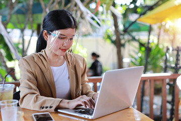 Young asian woman in casual dress with face shield and protect mask for healthcare, sitting in coffee shop and working on computer laptop and smartphone. New normal and social distancing concept