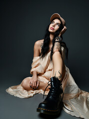 Sexy brunette woman with tattoo on arm and piercing on face in cap, evening maxi dress and massive...