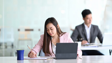 Young Asian businesswoman working with modern devices and sitting with her colleague in office.