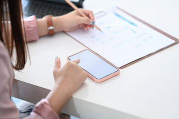 Close up view of businesswoman writing on paperwork and looking information on mobile phone.