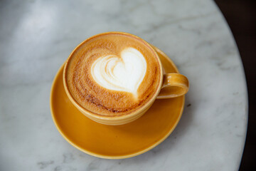 Trendy yellow cup of hot cappuccino on marble table background. Heart shape latte art for symbol of love