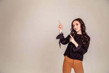 Fototapeta na wymiar beautiful slender young girl with black long hair on an isolated beige and gray background. woman in brown trousers and black blouse shows with forefinger hand